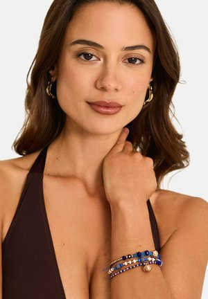 Jonge vrouw met golvend bruin haar, gouden oorringen en meerdere blauwe kralenarmbanden, gekleed in een donker haltertop.