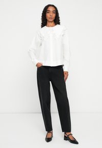 Donna in piedi contro un muro bianco, che indossa una blusa bianca con un ampio colletto di pizzo, jeans neri a vita alta e scarpe nere con tacco basso.