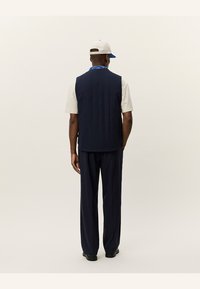 Navy padded vest with vertical seams, paired with a cream shirt and dark pants. Back view shows a casual look with a cap and bandana.