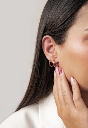 Nahaufnahme von einem rechten Ohr einer Frau mit zwei kleinen goldenen Creolen und einer Hand, die ihr Gesicht berührt, mit rosa Nägeln und weißen Punkten.