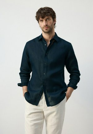 Uomo con capelli ricci castani indossa una camicia blu scuro abbottonata con le maniche arrotolate e pantaloni beige chiaro, in piedi davanti a uno sfondo semplice.