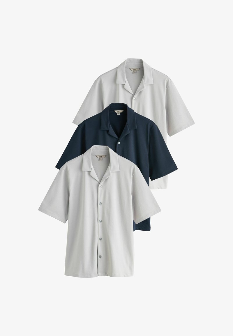 Trois chemises à manches courtes à boutons, en gris clair, bleu foncé et blanc cassé, disposées en chevauchement sur un fond blanc.