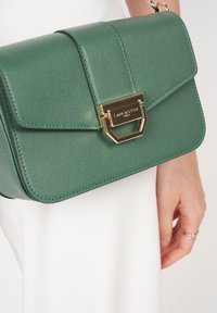 Grüne Leder-Schultertasche mit strukturierter Form, überklappbarem Deckel, goldenen Beschlägen und geprägtem Logodetail auf der Vorderseite.