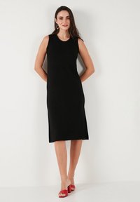 Black sleeveless dress with a straight cut, side slits, and smooth texture. Worn with red heeled sandals, against a neutral background.