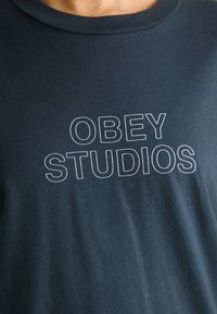 Tamno plava pamučna majica s okruglim izrezom; sadrži odvažne, bijele natpise "OBEY STUDIOS" ispisane na prsima. Mekana tekstura.