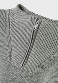 Primer plano de un cuello de suéter de punto gris con estrías, con una cremallera metálica parcialmente desabrochada.