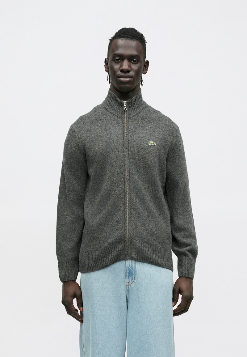 Šedý sveter na zips s rebrovaným golierom a manžetami. Zips vedie pozdĺž prednej časti, na ľavej strane je malý zelený logotyp. Klasická pletená štruktúra.
