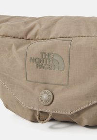 Beige szövet táska texturált felülettel, kiemelkedő logóval ellátott folttal és kerek fedélzárral, amely patenttal záródik.