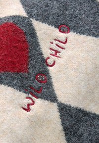 Stickad ullfilt med ett diamantmönster i kräm, grått och rött, med broderad text "WIL O CHI LO" i röda trådar. Mjuk textur.