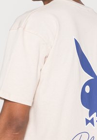 Beige kortärmad skjorta på person som visar en blå stiliserad kaninhuvudlogotyp med fluga på ryggen.