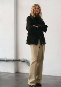 Giacca in velluto a coste nera con cappuccio foderato in pelliccia, design a strati. Abbinata a pantaloni beige a gamba larga e scarpe nere a punta.