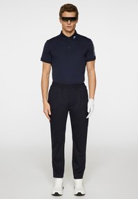 Marinblå polo skjorta med krage och knapplist, i kombination med mörka byxor med elastisk midja. Vita tennisskor och en vit handske.