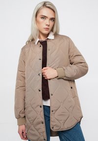 Quiltsēta bēša jaka ar spiedpogu aizdari, ribotām piedurknēm un brīvu piegriezumu. Uzvilkta virs bordo mikincera un balta krekla ar apkakli.