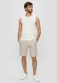 Weißes ärmelloses Shirt, kombiniert mit hellbeigen Shorts mit zwei vorderen Taschen. Abgerundet mit weißen Sneakers vor einem neutralen Hintergrund.