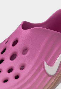 Κοντινό πλάνο ενός ροζ παπουτσιού τύπου slip-on από αφρώδες υλικό με οπές αερισμού και λογότυπο Nike Swoosh σε λευκό χρώμα στη πλευρά.