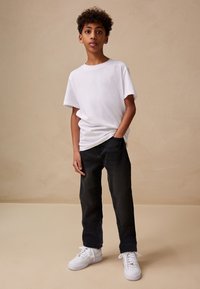 T-shirt blanc à manches courtes, jean noir à jambes droites, baskets blanches. Modèle debout, une main dans la poche, sur un fond neutre.