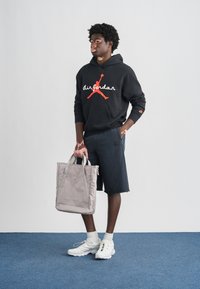 Μαύρο φούτερ με κουκούλα με λευκό λογότυπο «Air Jordan», σκούρο σορτς και λευκά αθλητικά παπούτσια. Κρατάει μια τσάντα tote ανοιχτό γκρι με γκρι χερούλια.