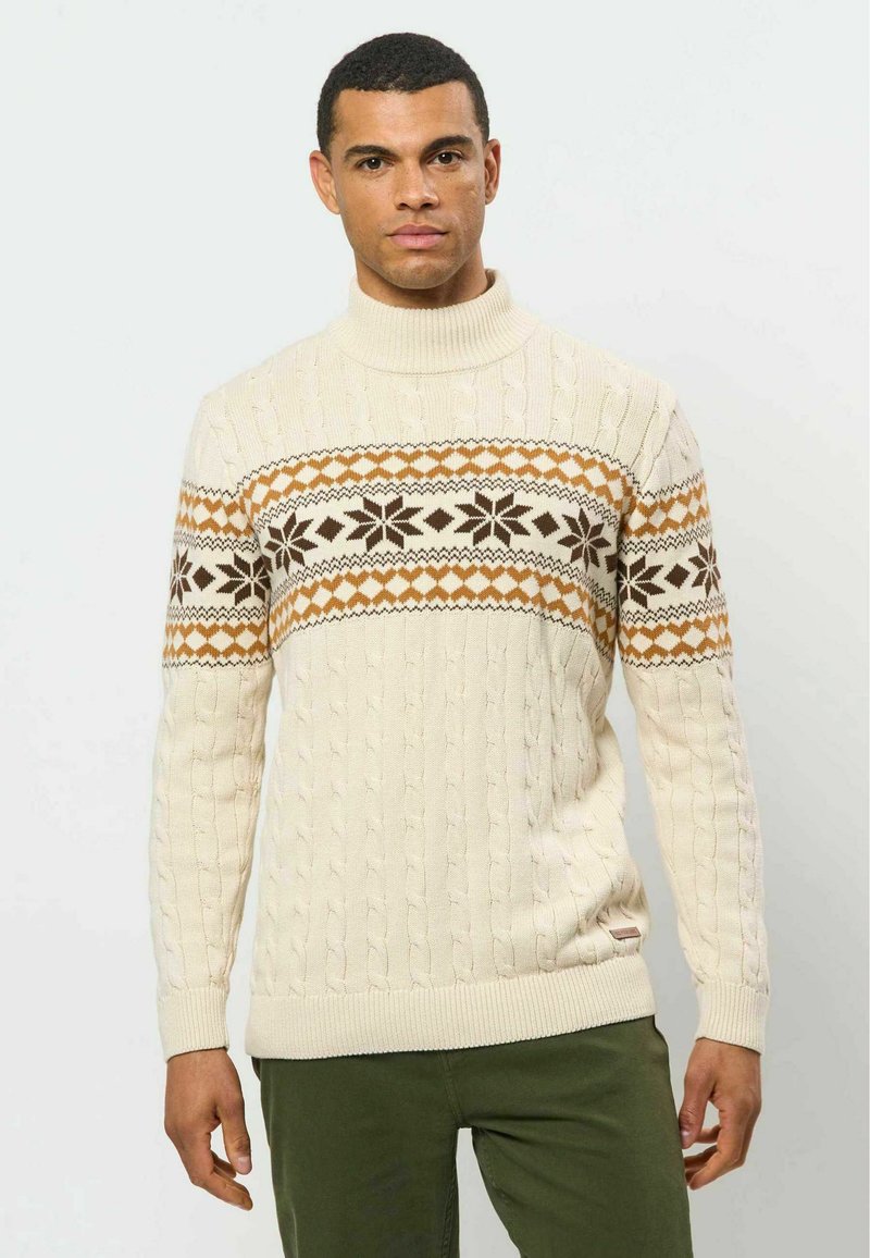 Homme portant un pull en maille torsadée crème avec un motif de flocons de neige et de losanges marron et orange sur la poitrine et un pantalon vert olive.