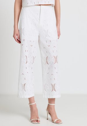 Woman wearing white high-waisted wide-leg pants with a floral eyelet pattern and white ankle-strap high-heeled sandals.
