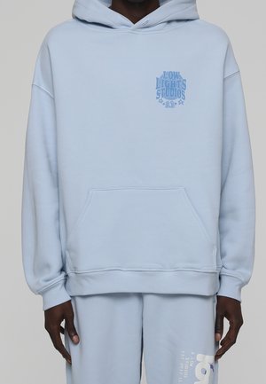 Personne portant un sweat à capuche bleu clair et un pantalon de survêtement assorti avec le texte "Low Lights Studios 23" sur la poitrine et un texte imprimé sur le pantalon.
