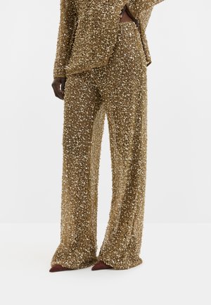 Pantalon long doré à sequins porté avec un haut assorti, assorti de chaussures pointues marron foncé, présenté sur une personne debout sur un sol blanc.
