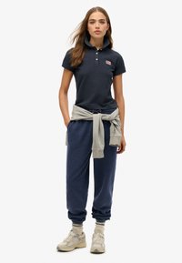 Marineblå polo-shirt med krave og logo, sammen med marineblå joggers og en lys grå sweatshirt bundet om taljen. Casual fodtøj fuldender looket.