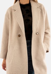 Abrigo de mezcla de lana beige claro con cuello con muescas, dos botones y bolsillos laterales. Textura suave y corte relajado, combinado con una blusa negra.