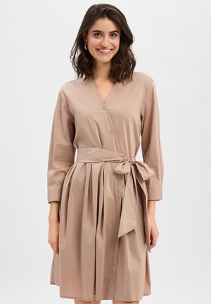 Kobieta o ciemnych włosach do ramion uśmiecha się, ubrana w beżową sukienkę z kopertowym dekoltem, wiązaną w talii, z długimi rękawami na jednolitym tle.