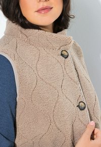 Gilet in pile beige e texture, con colletto alto e motivo a onde decorativo. Presenta bottoni neri e tessuto a maglia a contrasto sotto.