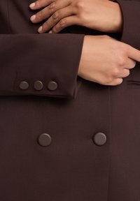 Brązowy płaszcz dwurzędowy z trzema matowymi brązowymi guzikami na rękawie, charakteryzujący się gładką fakturą i dopasowanym krojem.