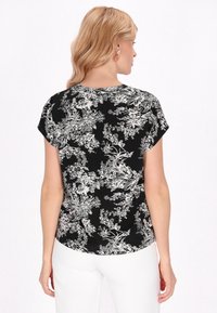 Camicia nera a maniche corte con motivo floreale bianco, dotata di orlo arrotondato e texture morbida. Indossata con pantaloni bianchi, vista da dietro.