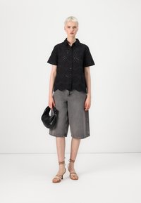 Maglietta nera a maniche corte con dettagli a occhio, orlo scallop, abbinata a pantaloni corti in denim grigi, borsa nera e sandali con cinturini color sahara.