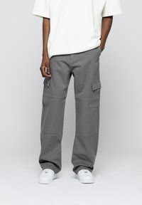 Pantalon cargo gris à coupe ample, avec poches plates sur les côtés, coutures renforcées et tissu résistant. Associé à des baskets blanches.