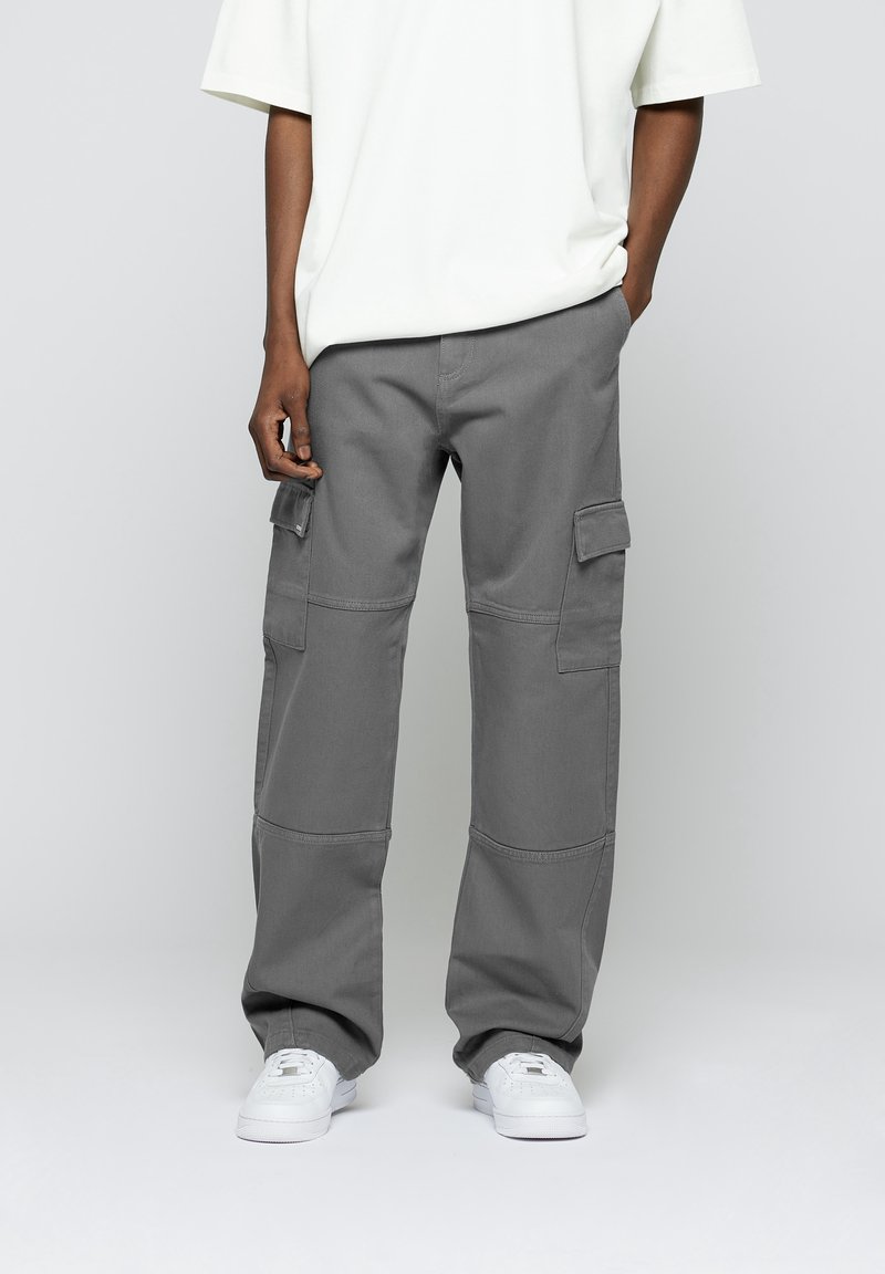 Pantalon cargo gris à coupe ample, avec poches plates sur les côtés, coutures renforcées et tissu résistant. Associé à des baskets blanches.