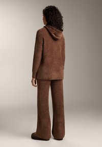 Hnedá mikina s kapucňou a rozšírené nohavice z mäkkej, textúrovanej látky; obsahujú kapucňu a voľný strih. Farba je bohatá, tmavohnedá.