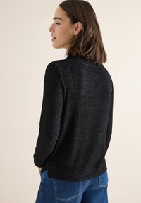 Černý textured svetr s dlouhými rukávy a zakulaceným výstřihem, kombinovaný s modrými džínami. Jednoduchý a moderní design, vhodný pro volnočasové nošení.