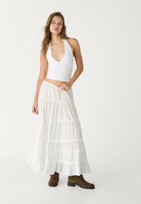Vit halterneck crop top och lång vit kjol med tierade volanger, kombinerad med bruna ankelboots. Slätt tyg med en flödig siluett.