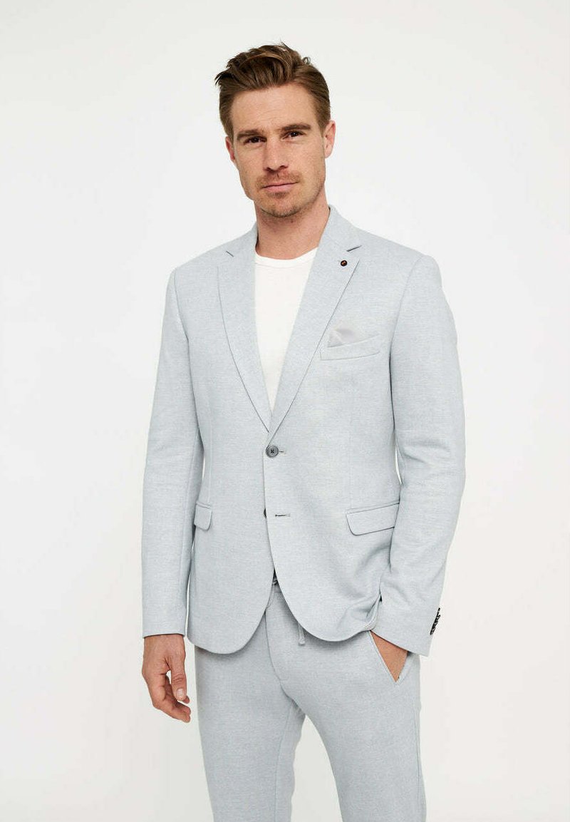 Lichtgrijs pak met een notch lapel, enkele knoopsluiting en een getailleerde pasvorm. Beschikt over een borstzak en zijzakken, gecombineerd met een effen wit overhemd.