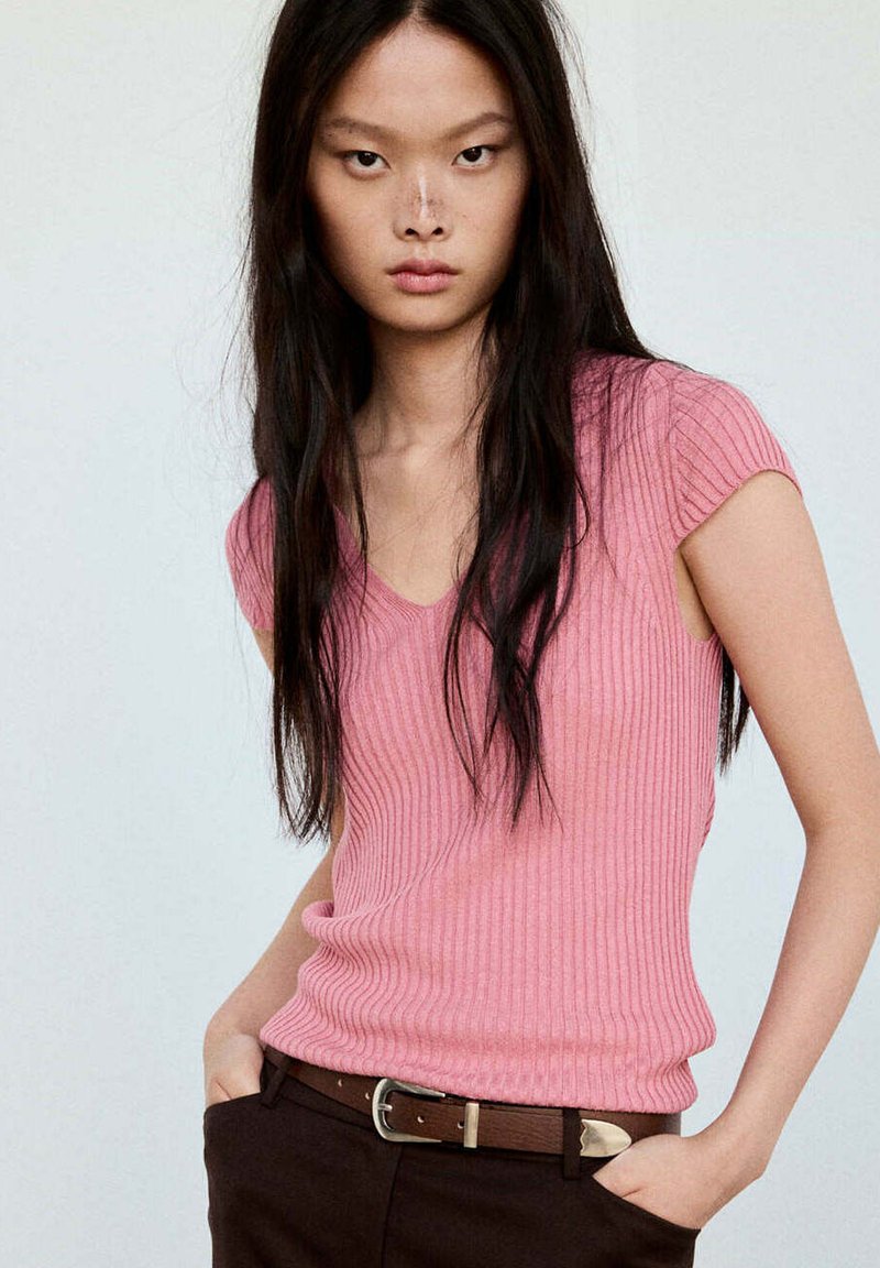 Jeune femme aux longs cheveux foncés portant un haut rose côtelé à manches courtes et un pantalon foncé avec une ceinture marron sur un fond uni.