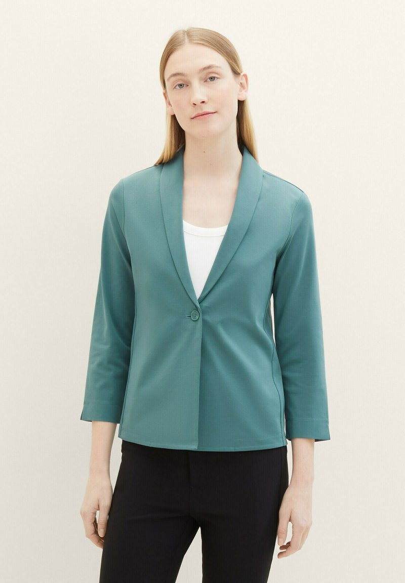 Vrouw met steil blond haar, gekleed in een teal blazer over een witte top en zwarte broek, staand tegen een effen lichte achtergrond.