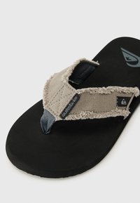 Men's flip-flop featuring a light beige fabric strap with frayed edges, dark gray accents, and a black rubber sole. Design includes a logo detail.