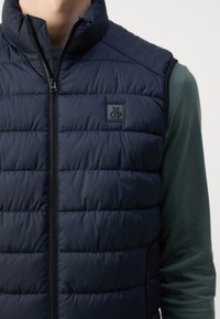 Marineblaues gepolstertes Weste mit hohem Kragen, Reißverschluss und Logo-Patch. Merkmale sind gesteppten Linien und ein schlankes, leichtes Design.