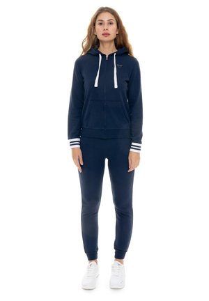 Une femme porte un sweat à capuche bleu marine zippé, assorti à un pantalon de jogging bordé de blanc, ainsi que des baskets blanches, sur un fond blanc uni.
