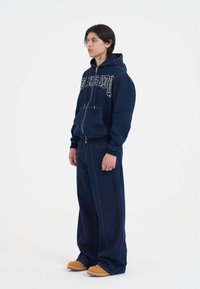 Marineblaue Denim-Hoodie mit weißem Schriftzug-Logo, kombiniert mit weit geschnittenen Denim-Hosen und beigen Schuhen. Verfügt über einen Reißverschluss und Taschen.