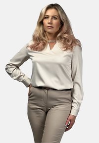 Beżowa bluzka z długim rękawem i dekoltem w serek, o gładkiej fakturze, w połączeniu z dopasowanymi beżowymi spodniami i czarnym paskiem.