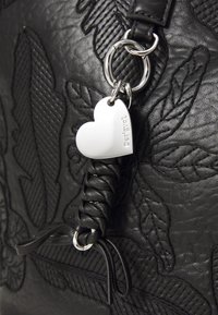 Bolso de mano de cuero negro con patrones de hojas grabados, detalle de asa trenzada y una etiqueta en forma de corazón plateada con el nombre de la marca.