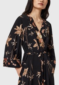 Vestido negro de flores con mangas largas, que presenta patrones de flores en beige y marrón, un escote en V y una cintura elástica. Incluye bolsillos.