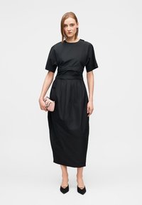 Black dress with short sleeves, cinched waist, and a midi length. Model holds a small pink handbag, paired with black pointed-toe heels.
