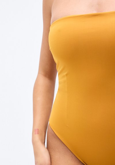 Ολόσωμο μαγιό χωρίς τιράντες σε ζεστό μουσταρδί χρώμα. Διαθέτει λεία ύφη, εφαρμοστή γραμμή και ψηλό κόψιμο στο πόδι.