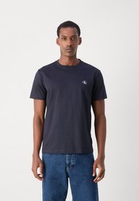 Marineblå bomulds t-shirt med rund hals og korte ærmer, der har et lille hvidt logo på venstre bryst. Afslappet pasform og glat tekstur.